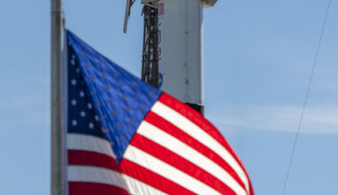 A close-up view of a SpaceX Crew Dragon spacecraft mounted atop a Falcon 9 rocket on the launch pad. The capsule and trunk are connected to ground support hardware, with NASA and mission insignia visible on the rocket’s exterior against a bright blue sky. in the foreground in the American flag.