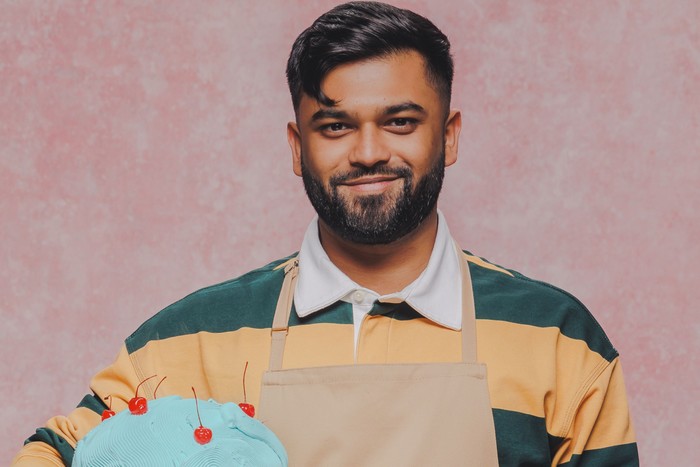7 A man stood smiling in front of a pastel pink background, holding a cake under one arm.