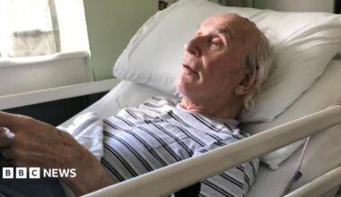 John Russell is an older man. he is wearing a black and white stripy T'shirt and lying back in a hospital bed.