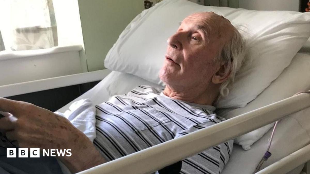 John Russell is an older man. he is wearing a black and white stripy T'shirt and lying back in a hospital bed.