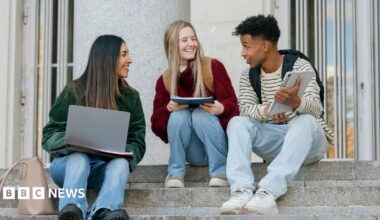 Three university students sitting on concrete stairs, talking and smiling while looking at a digital tablet and laptop. There are two women to the left both looking at a male student to the right.