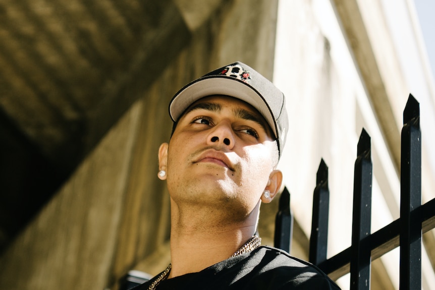Yung Milla, wearing a cap, leans against a wall looking up.