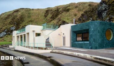 Tarlair Pool Pavilion, a restored Art Deco building beside the seawater pool, featuring cream-coloured walls, pale green railings, rooftop terraces, and a modern dark green extension, set against a rugged coastal hillside.