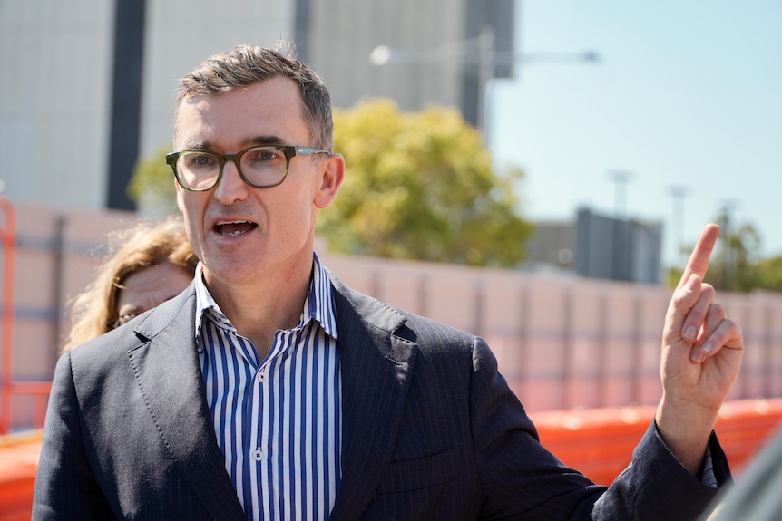 John Carey speaking, wearing striped shirt and black blazer, gesturing by holding up index finger