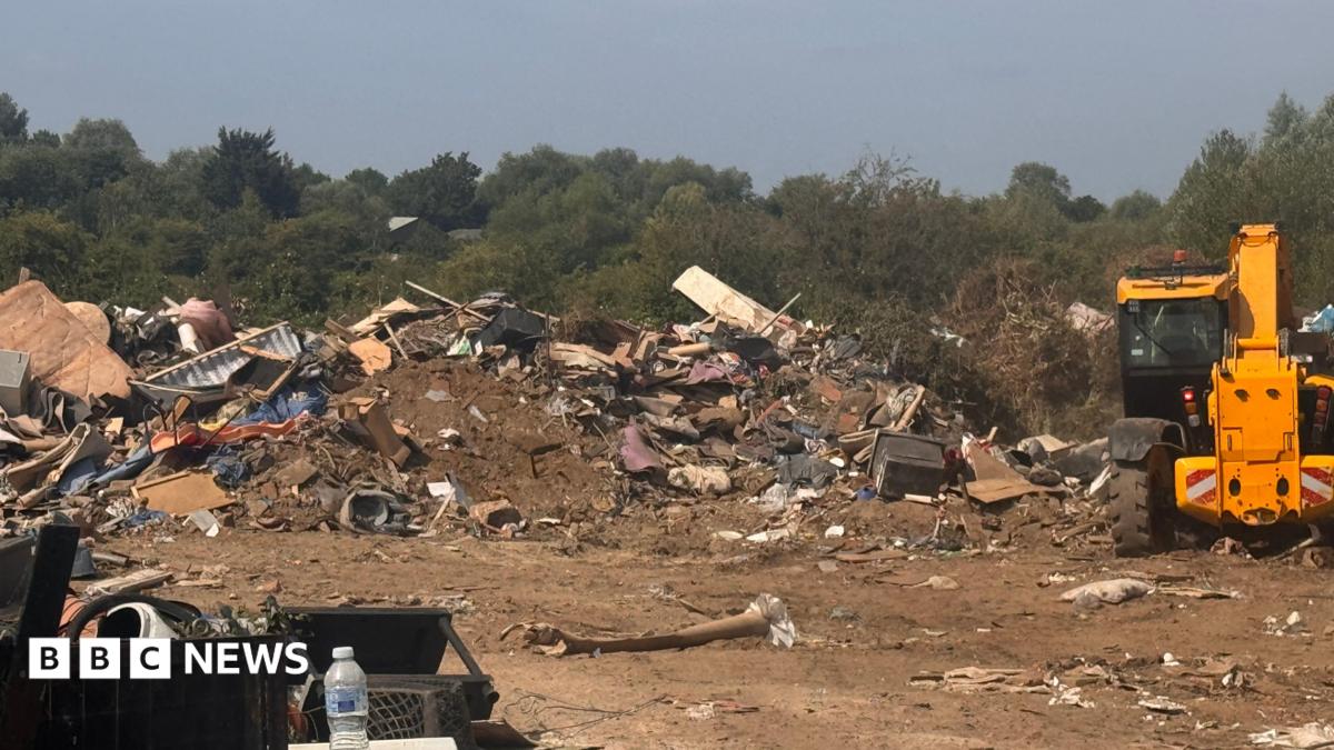 A site with a big pile of dumped waste. There is a yellow digger next to the pile and lots of trees in the background.