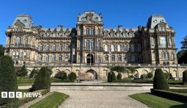 A large building in the French Renaissance style of architecture - four storeys with large windows, projecting bays, and mansard roofing. Stairs on either side lead up the the central entrance. In front is a formal garden with a wide gravel path, patches of lawn, and clips shrubbery.