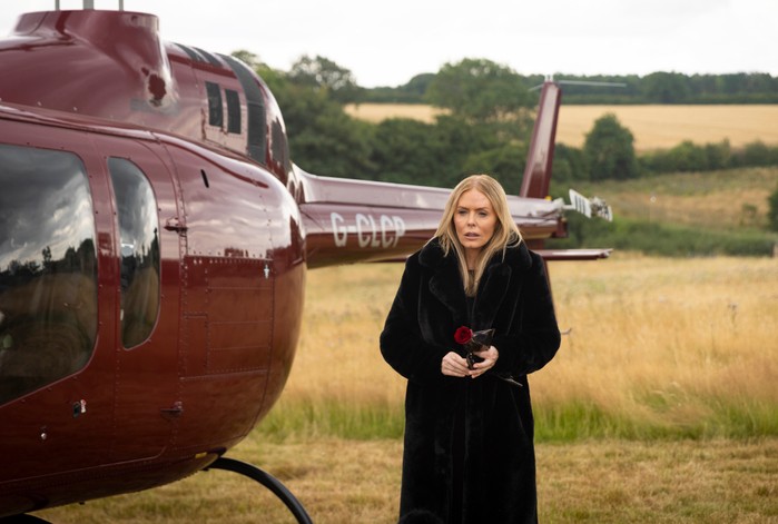 Sadie steps away from a red plane, wearing a long black coat and holding a red rose in Emmerdale