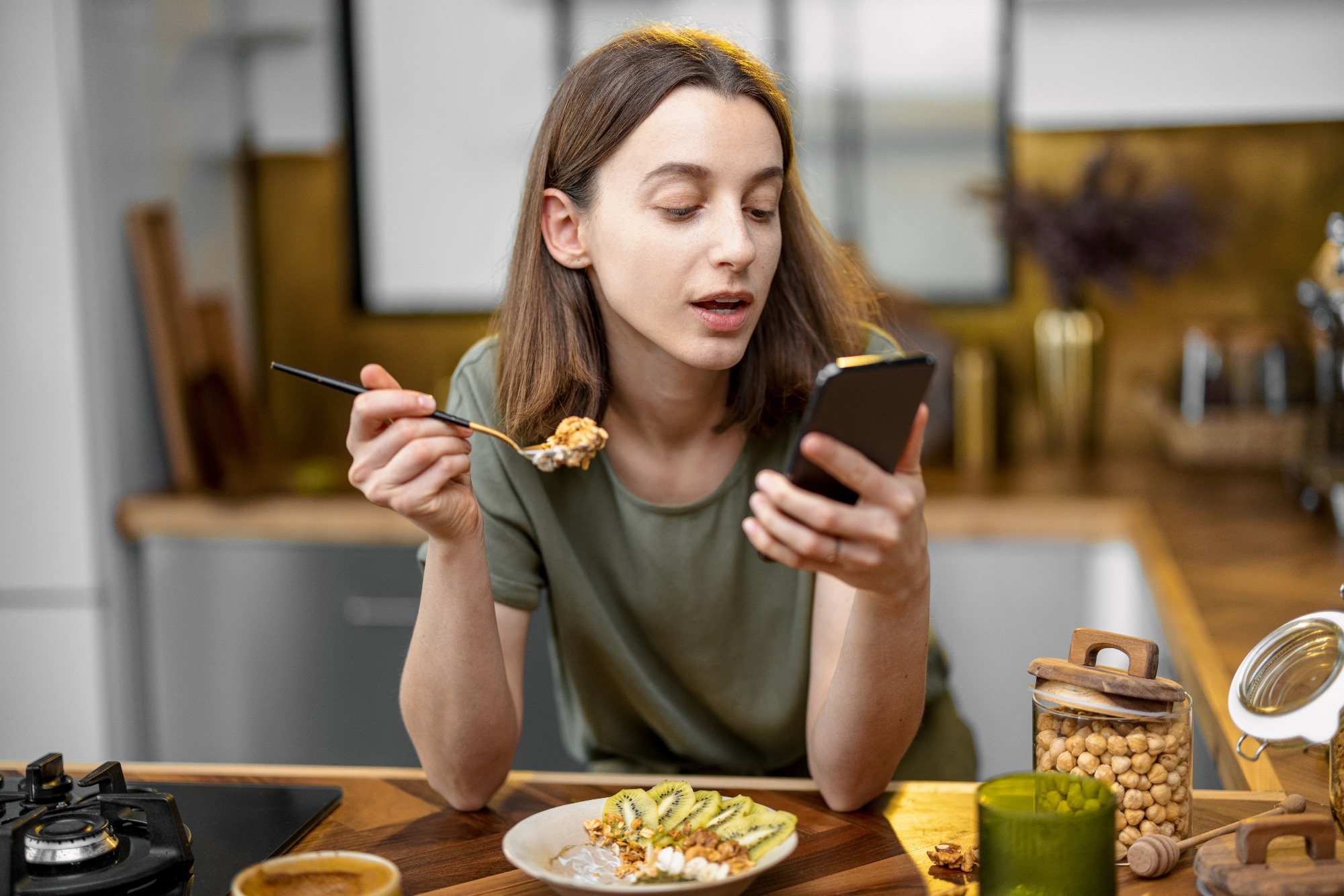 Woman in casual green t-shirt holds a spoon with granola in her hand and reads on the phone during a morning time on the kitchen at home.