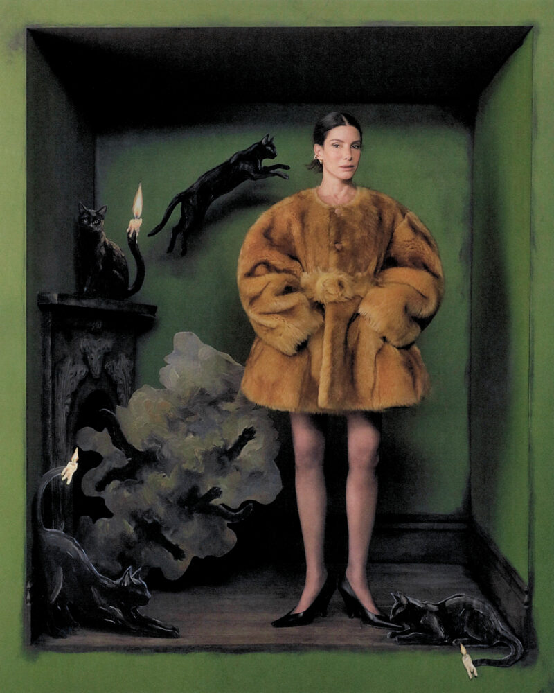 Sandra Bullock stands before a green wall with her feet turned outward and hands in the pockets of a fur coat. She is surrounded by cut-outs of black cats.