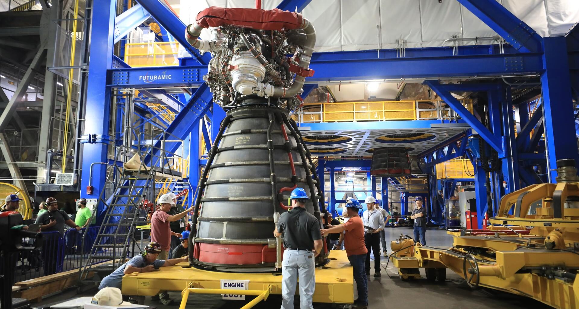 A rocket engine sits on a yellow transport platform inside a large industrial facility. Several technicians wearing hard hats and safety gear stand around the engine, inspecting and guiding it. The engine’s bell nozzle faces downward, and complex piping and hardware are visible at the top. Bright blue structural frames and elevated yellow walkways surround the work area, with additional equipment and machinery in the background.