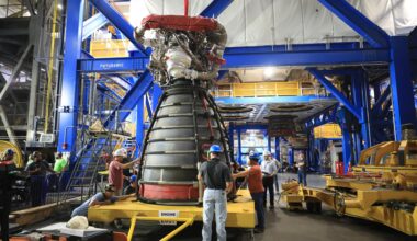 A rocket engine sits on a yellow transport platform inside a large industrial facility. Several technicians wearing hard hats and safety gear stand around the engine, inspecting and guiding it. The engine’s bell nozzle faces downward, and complex piping and hardware are visible at the top. Bright blue structural frames and elevated yellow walkways surround the work area, with additional equipment and machinery in the background.