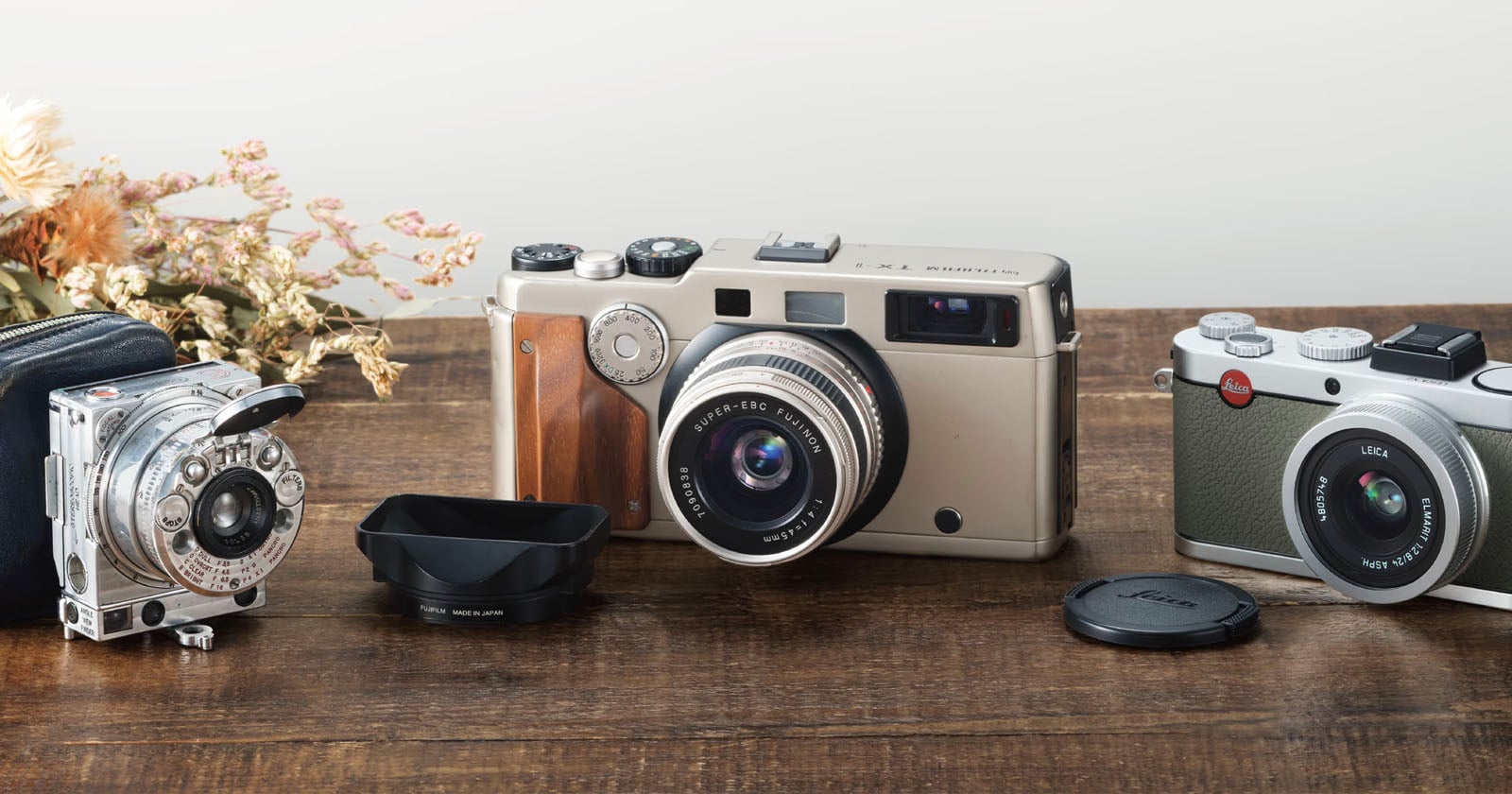 Three vintage cameras and a lens hood sit on a wooden table next to a small bouquet of dried flowers.