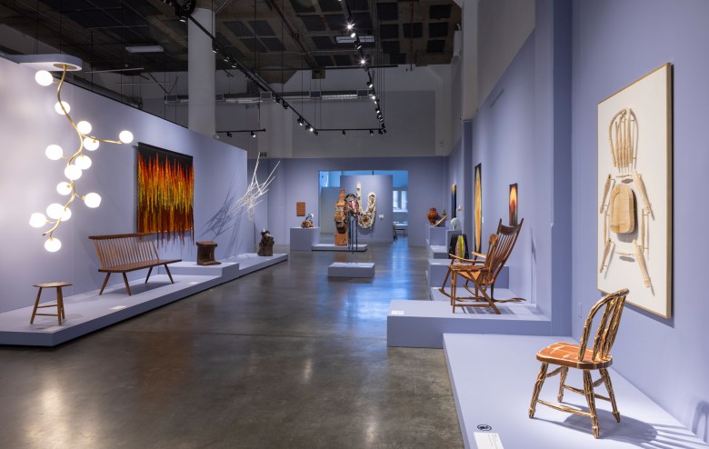 Contemporary art gallery with modern and wooden furniture pieces, sculptures, and abstract paintings displayed on light purple walls and platforms.