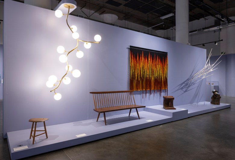 A modern gallery display features a branch-like light fixture, wooden bench and stool, colorful wall textile, abstract sculpture, and a white angular installation against a pale lavender wall.