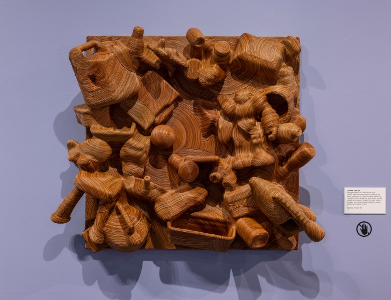 Layered wooden sculpture of various abstract and recognizable objects densely packed on a square panel, mounted on a light purple wall with a small information placard to the right.