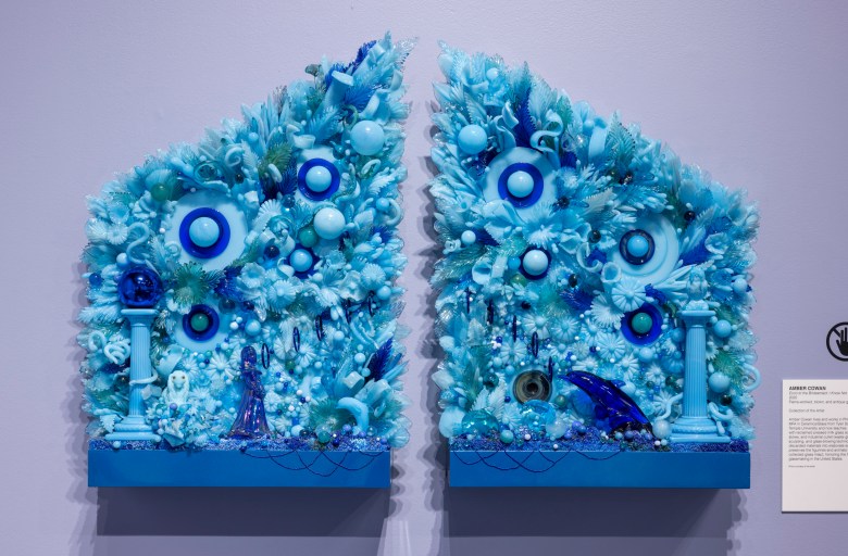 Mixed-media wall sculpture in two parts, featuring various blue objects including seashells, toys, and artificial plants, arranged densely against a lavender wall.