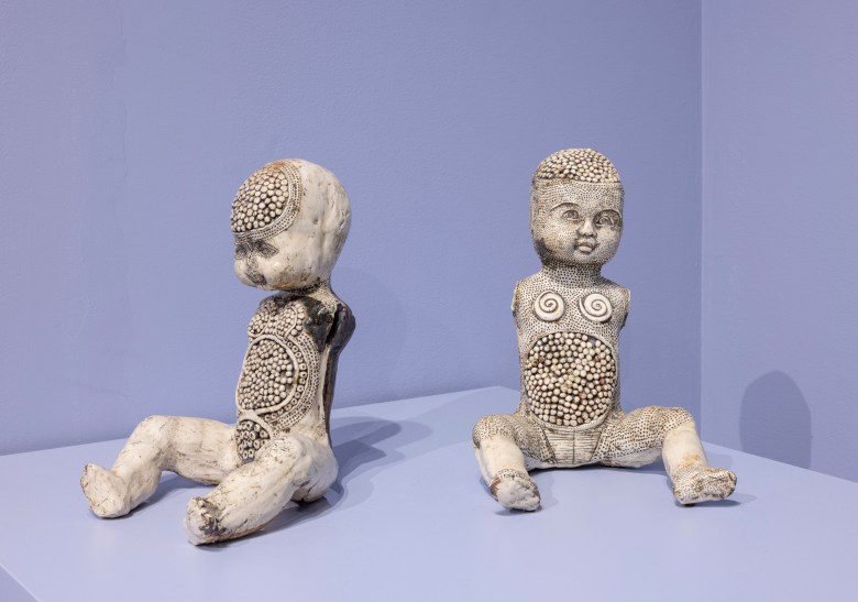 Two seated sculptures resembling babies, decorated with circular patterns and small shells, displayed against a light purple wall.