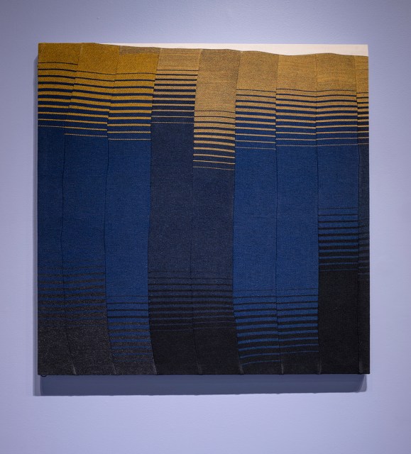 Textile artwork with warped horizontal lines in shades of yellow, blue, and black, forming a grid-like pattern on a square canvas against a light purple wall.