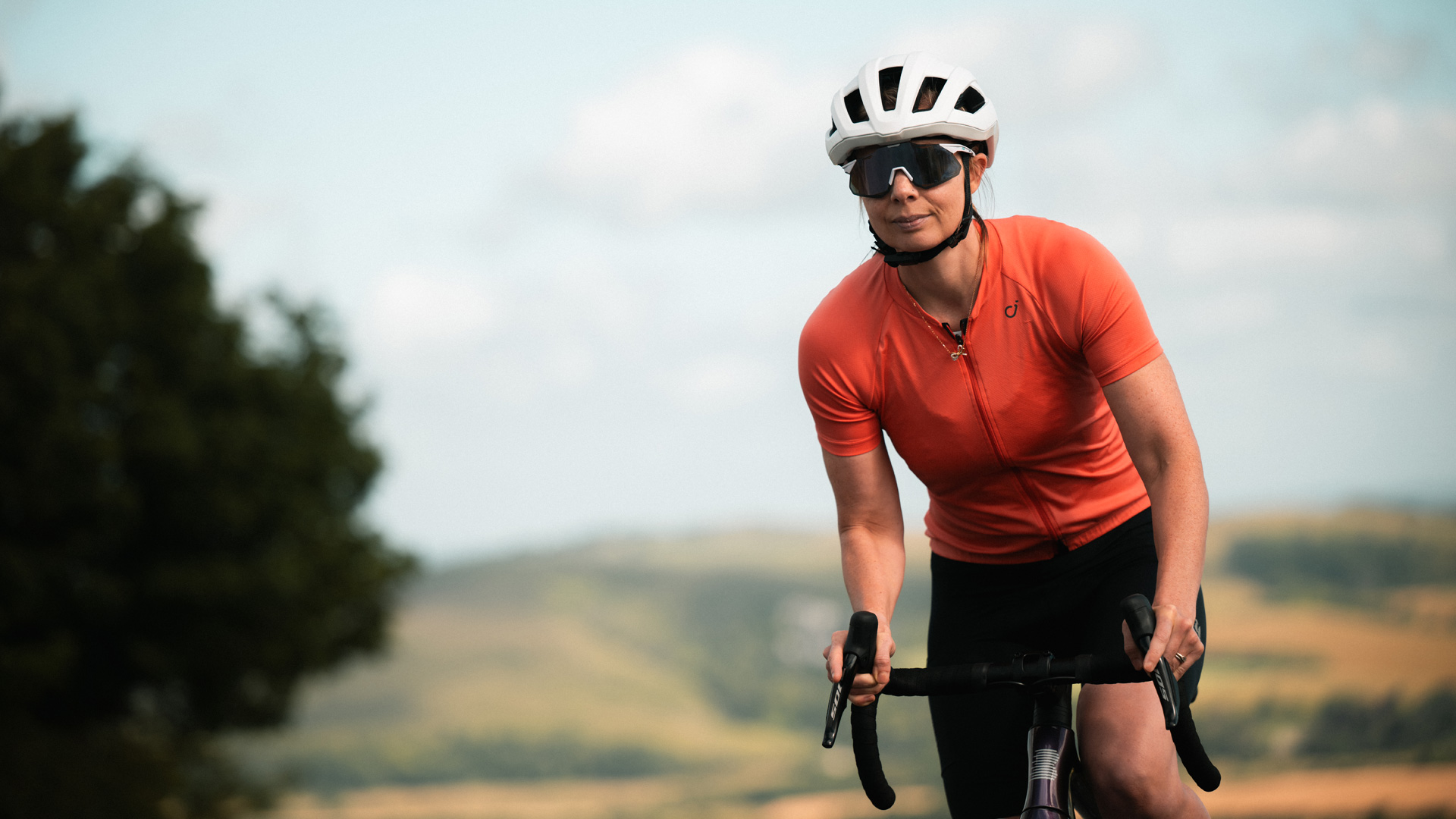 A cyclist rides on the hoods up a climb with a Sussex background