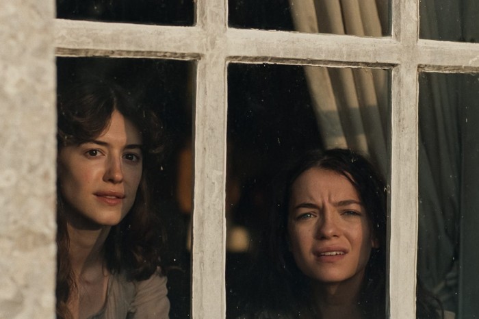 Daisy Edgar-Jones (left) and Esmé Creed-Miles star as sisters Elinor and Marianne. The differences between the pair are a key focus of the story | Credit: © 2026 FOCUS FEATURES Film still viewed through a white-framed window with multiple panes, looking in at two young women peering out from a dim interior lit warmly behind them. The woman on the left has dark wavy hair and a calm, watchful expression; the woman on the right has darker hair, a furrowed brow, and an anxious or startled look, mouth slightly open