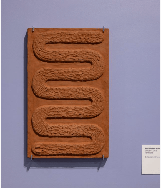 Rectangular terracotta sculpture with a raised, winding, textured path resembling a maze, mounted on a light purple wall with a label displayed to the right.