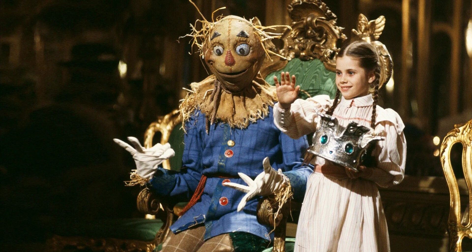 A still from Return to Oz showing Dorothy, in a striped dress and plaited hair, holding a jewelled silver crown beside the Scarecrow on an ornate gilded throne.