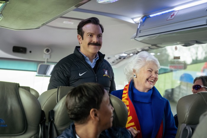 Ted_Lasso_Photo_040906 Jason Sudeikis and Annette Badland in Ted Lasso, stood on a coach, smiling.