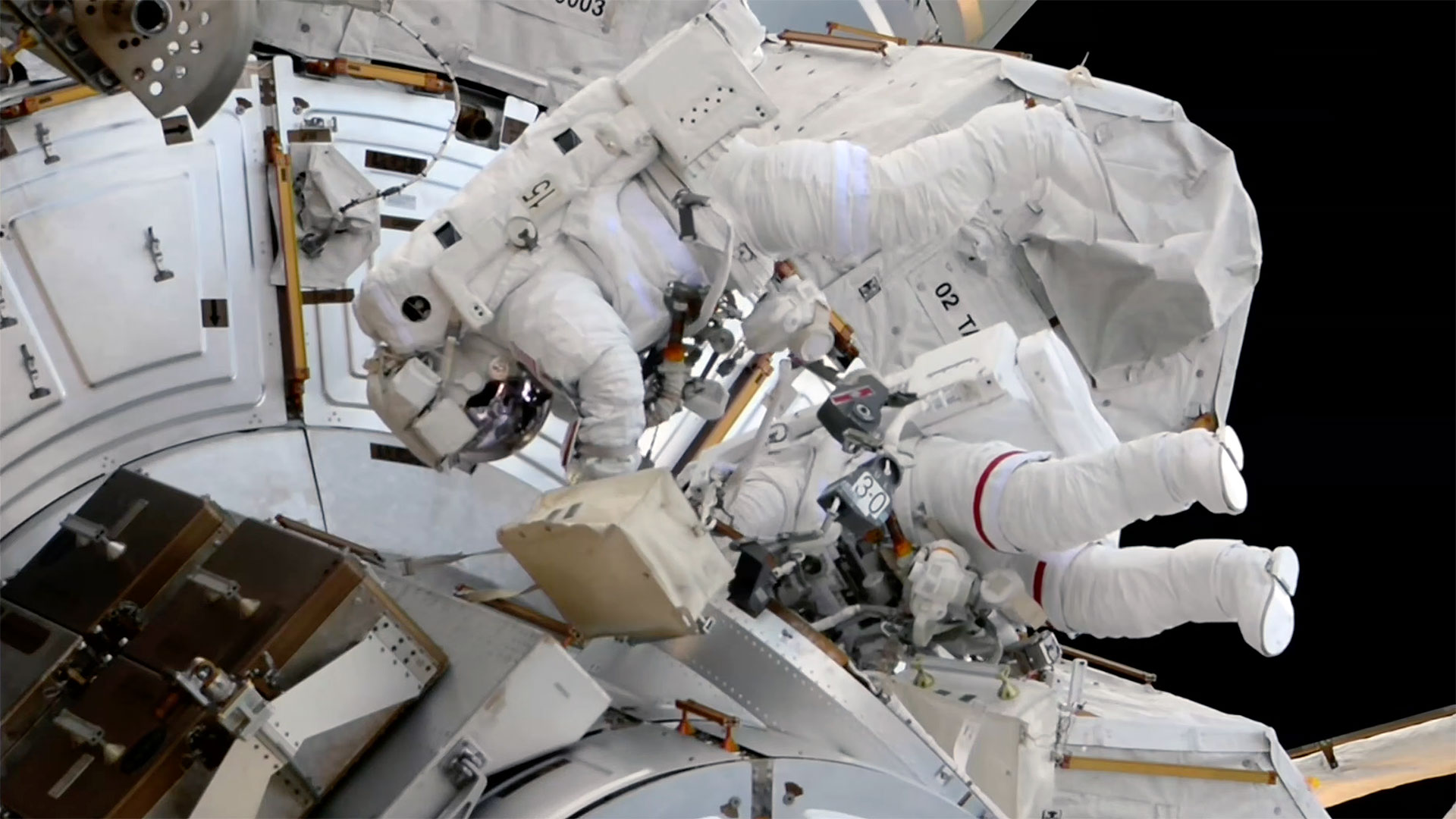 two astronauts in white spacesuits are seen outside a space station during a spacewalk
