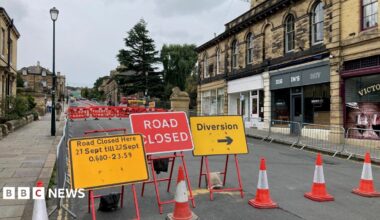 Yellow and red road signs showing road closure times and pointing to a diversion. There are red and whites barriers and traffic cones closing off the road.