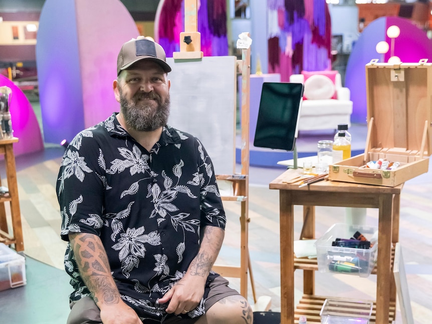 A man with a cap and beard sits next to a table with art supplies on it. He smiles at the camera
