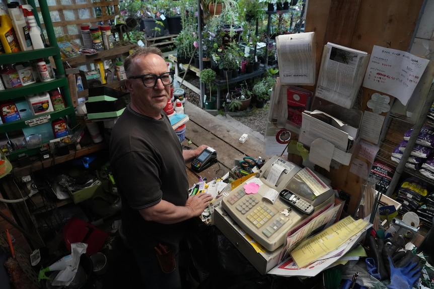 A man wearing classes stands behind a cash register inside a nursery.