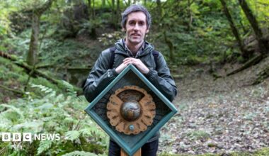 Artist Daniel Weatheritt posing with one of his sculptures at Stubsgill Beck, Distington. He has short salt and pepper hair and wears a dark green waterproof jacket and jeans. He is posing in the woods, standing behind his oak sculpture. The sculpture is an oak cog wheel inserted in a diamond shape frame.