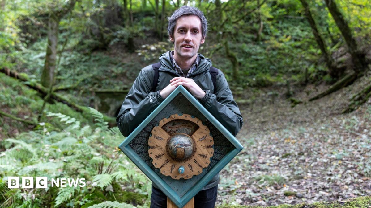 Artist Daniel Weatheritt posing with one of his sculptures at Stubsgill Beck, Distington. He has short salt and pepper hair and wears a dark green waterproof jacket and jeans. He is posing in the woods, standing behind his oak sculpture. The sculpture is an oak cog wheel inserted in a diamond shape frame.