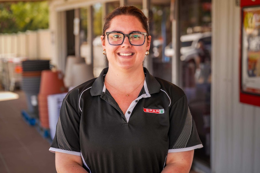 A woman with brown hair and glasses and wearing a uniform smiles out the front of a grocery store.