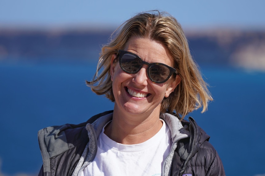 A woman smiling at the camera, wearing sunglasses, and standing in front of water.