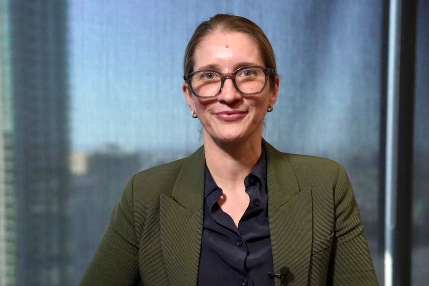 A woman wearing glasses with her hair pulled back smiles with her mouth closed she is wearing a black shirt and green blazer.