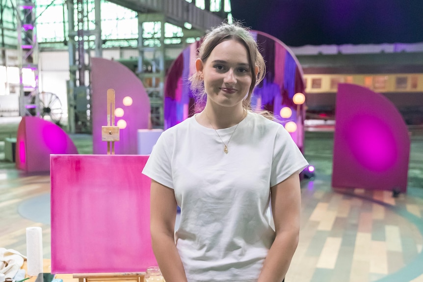 A young woman with brown hair smiles at the camera. She wears a white t-shirt. Behind her are pink shapes on a TV set