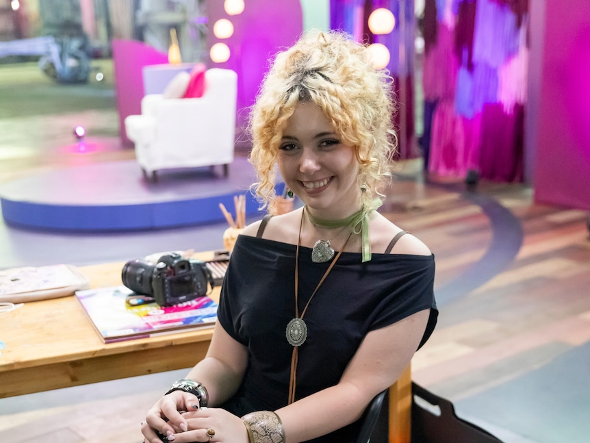 A young woman with curly blonde hair tied back smiles at the camera. A desk with a camera is behind her