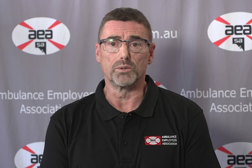 Paul Ekkelboom wears glasses as he stands in front of a grey ambulance union banner