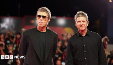 Liam and Noel Gallagher wearing black clothing stand on a red carpet with photographers stood behind them.