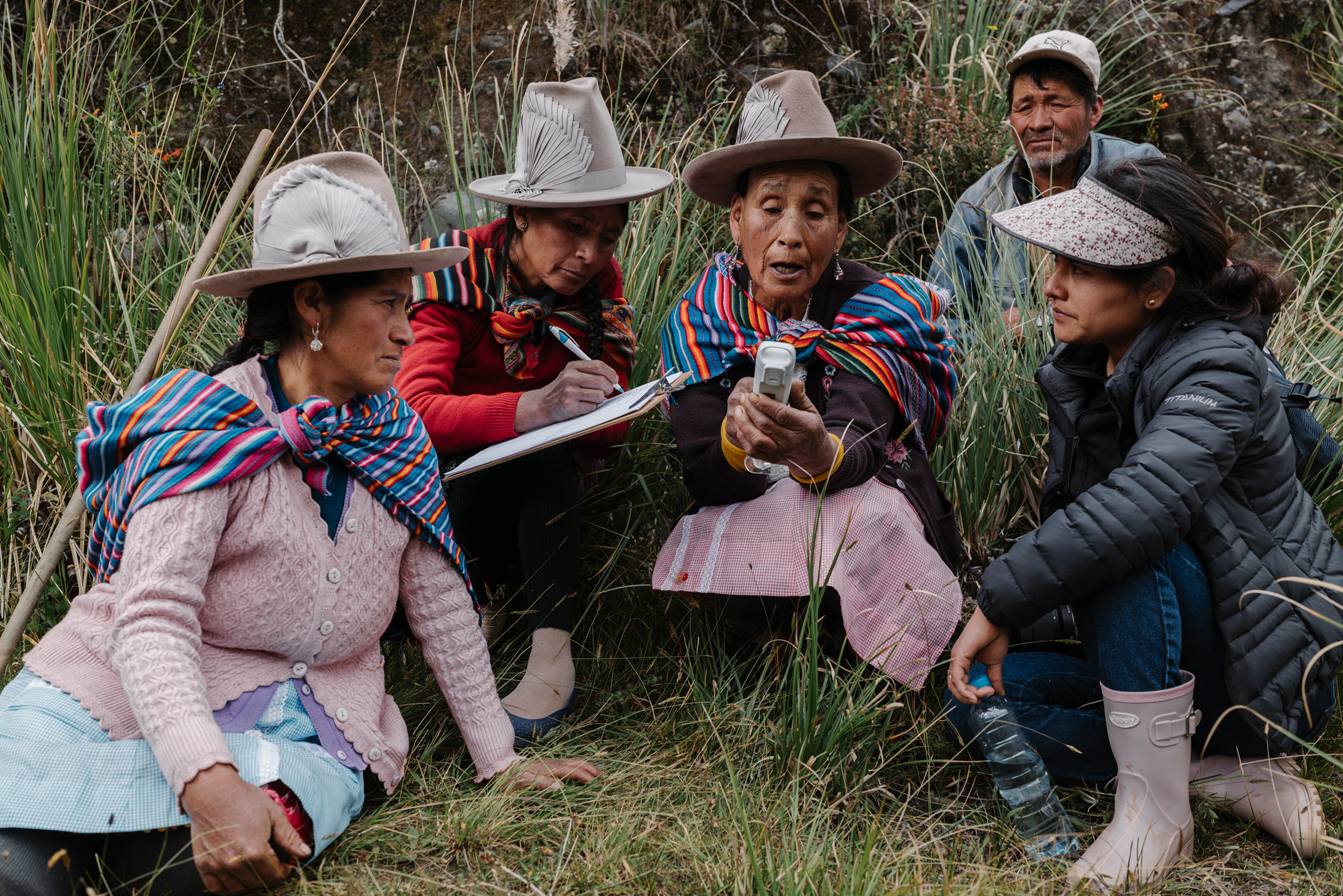 Five people, including women in traditional Andean clothing and hats, gather outdoors in tall grass, one woman holding a phone and another writing