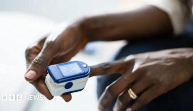 Close-up of a pulse oximeter clipped onto a person's finger. The blue-and-white device displays blood oxygen and pulse readings while the person's hands rest in their lap.