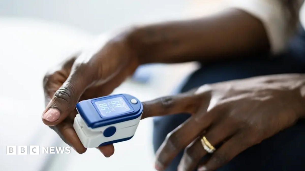 Close-up of a pulse oximeter clipped onto a person's finger. The blue-and-white device displays blood oxygen and pulse readings while the person's hands rest in their lap.