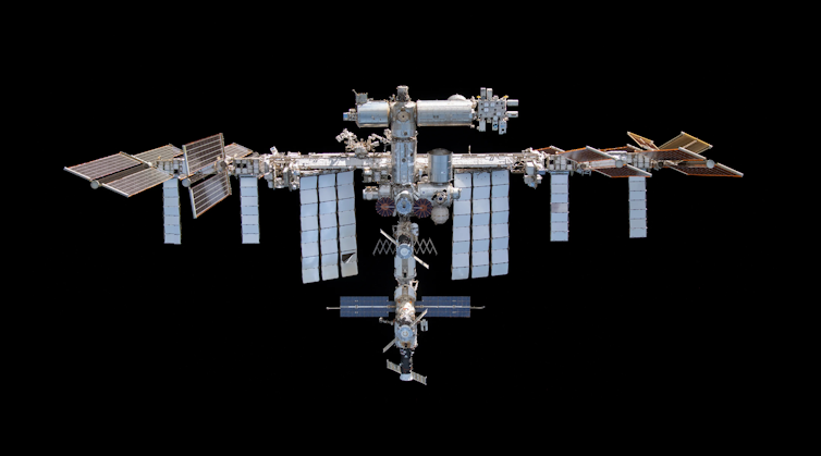 The International Space Station, which is a metal structure with solar panels coming off it, floating above Earth