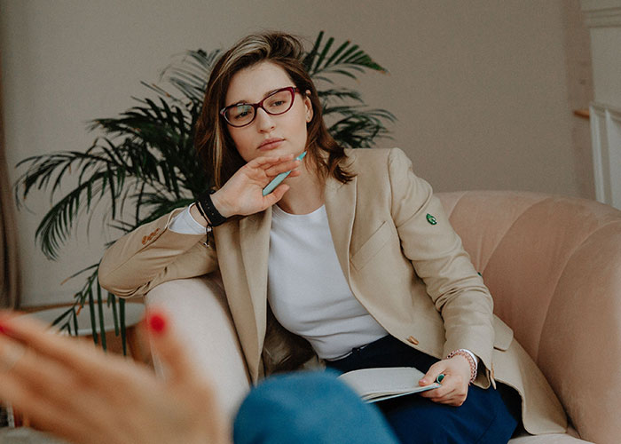 A woman in glasses and a blazer listens intently with a pen and notepad, discussing symptoms that doctors stop and stare at.