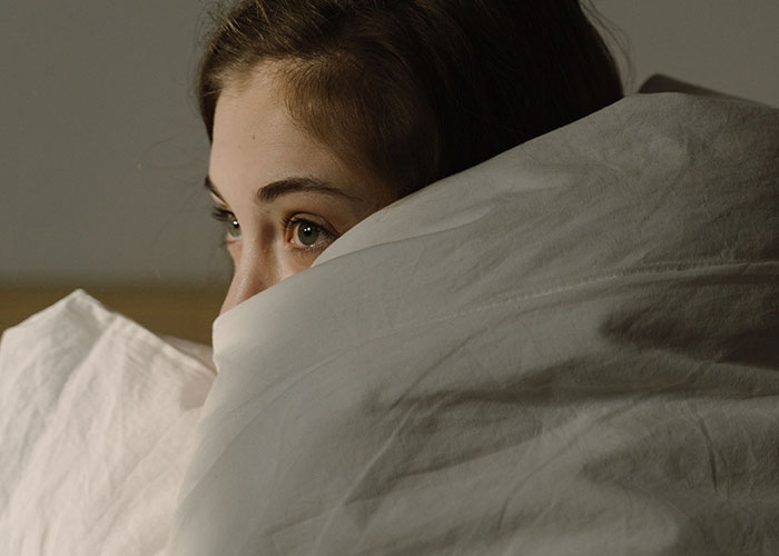 A woman peeks from under white sheets, eyes wide with worry over normal symptoms. Doctors are often perplexed.
