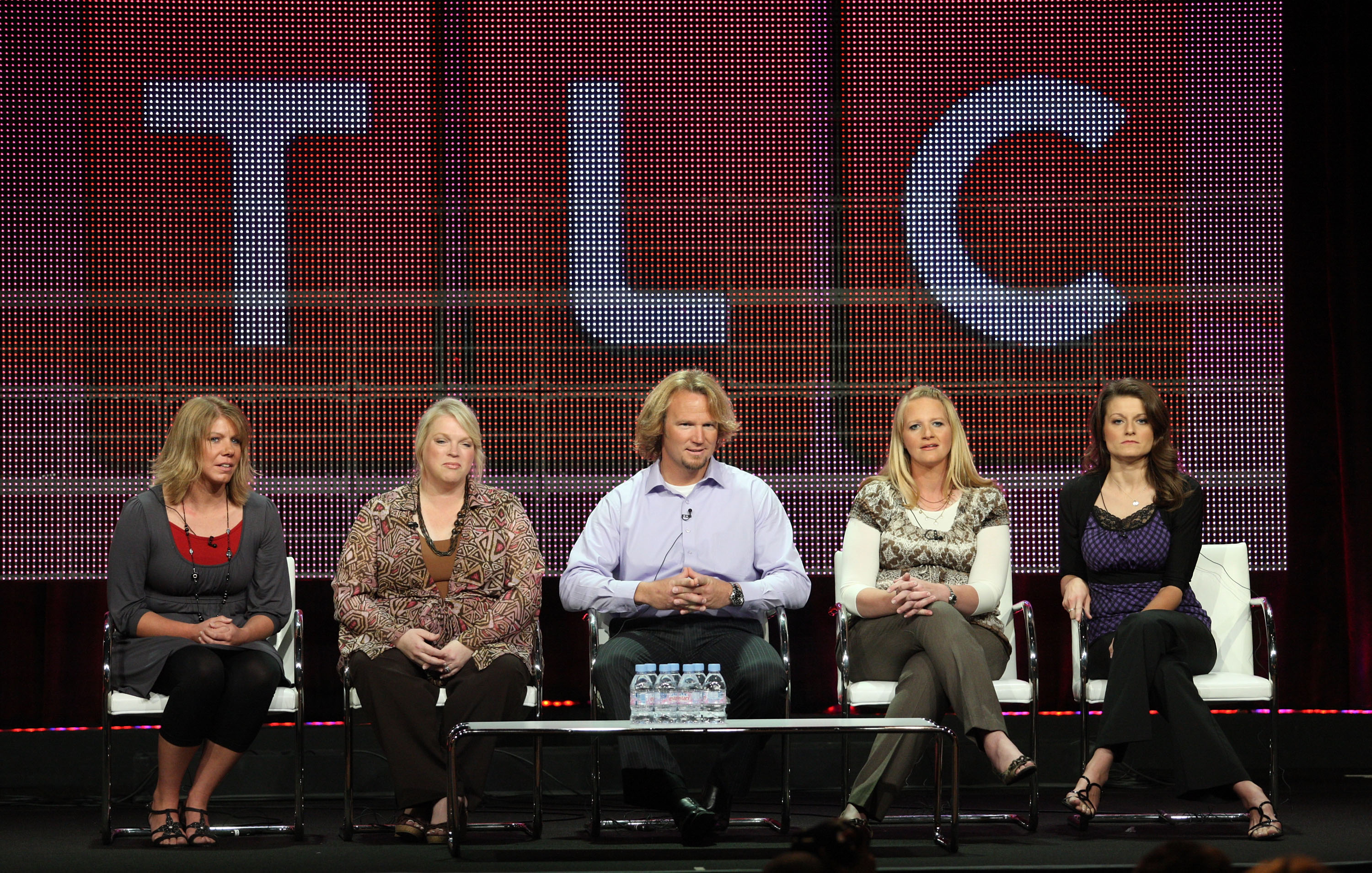 Five people seated in white chairs on a panel stage in front of a large TLC LED display.