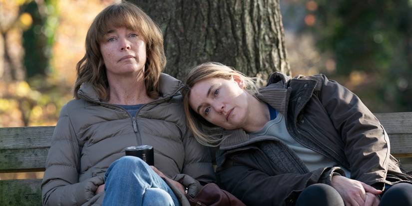 Julianne Nicholson sitting on a park bench with Kate Winslet's head on her shoulder in 'Mare of Easttown'.