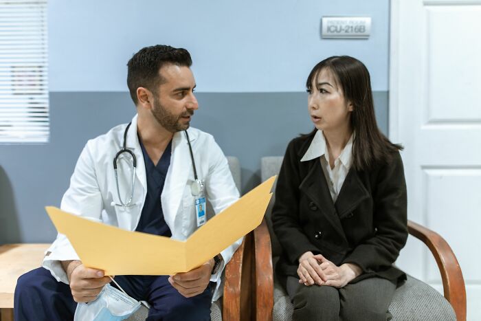 A medical worker in a lab coat discusses patient stories with a woman holding a yellow file.