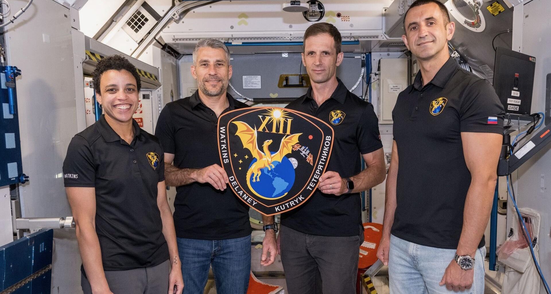 three men and one woman, all wearing black polo shirts, hold a plaque inside a mockup space module